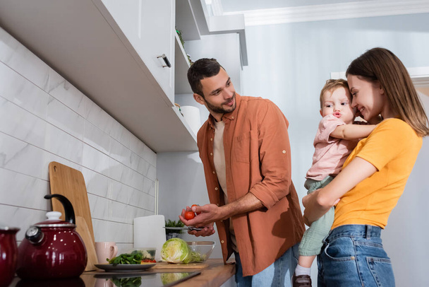 Positive mother hugging son while husband cooking salad in kitchen  - Photo, image