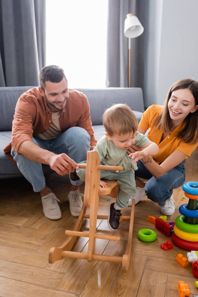 Happy parents holding kid near rocking horse and toys at home  - Foto, afbeelding