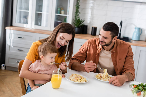 Smiling woman feeding pasta to son near husband and orange juice in kitchen  - Foto, afbeelding
