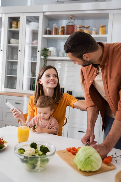 Smiling woman holding smartphone near husband cooking salad and son at home  - Photo, image