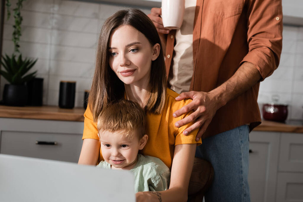 Hombre con taza abrazando esposa usando portátil cerca de hijo en casa  - Foto, Imagen