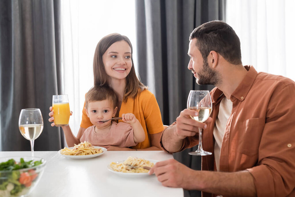 Uśmiechnięta kobieta trzyma sok pomarańczowy w pobliżu syna jedząc makaron i męża w domu  - Zdjęcie, obraz