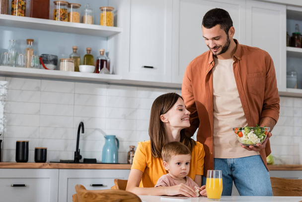 Smiling man holding salad near family and orange juice in kitchen  - Zdjęcie, obraz