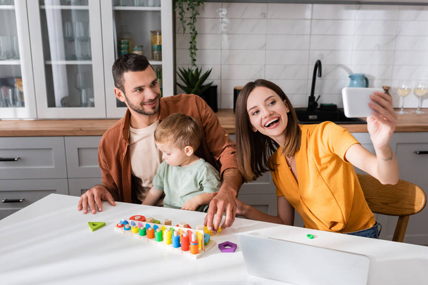 Femme heureuse prenant selfie avec la famille près de jeu éducatif et ordinateur portable à la maison  - Photo, image