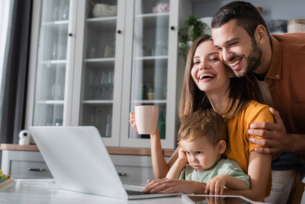 Positive man hugging wife with cup near kid and devices at home  - Foto, Imagen