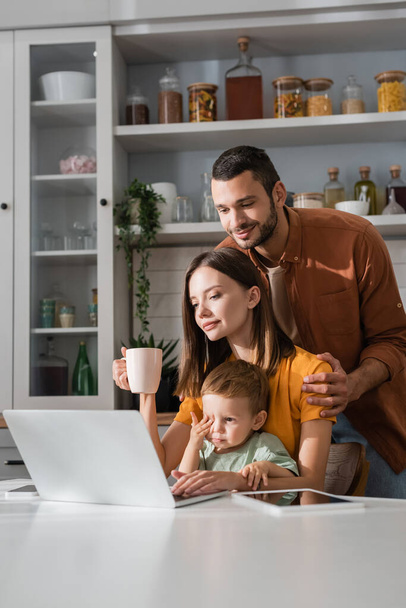 Man hugging wife with cup near gadgets and son at home  - Φωτογραφία, εικόνα