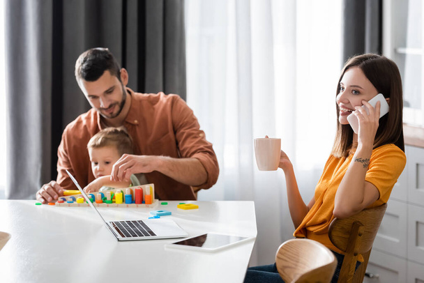 Smiling woman talking on cellphone and holding cup near laptop and family at home  - Foto, Imagen