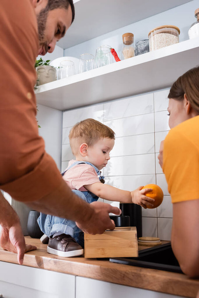 Ouders in de buurt van peuterkind met oranje op keukenblad  - Foto, afbeelding