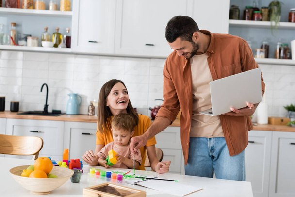 Man holding laptop and drawing near wife and son in kitchen  - Foto, immagini