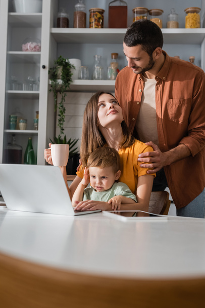Young man hugging wife with cup near son and gadgets at home  - Foto, Imagen