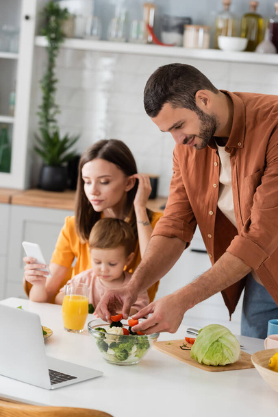 Man koken salade in de buurt van laptop en wazig gezin in de keuken  - Foto, afbeelding