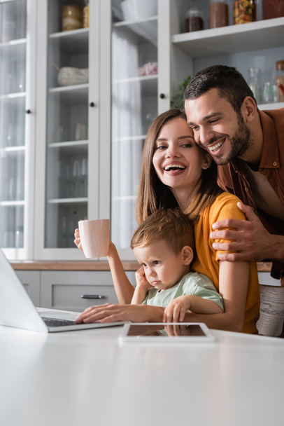 Happy man hugging wife with cup near son and gadgets at home  - Foto, Imagen