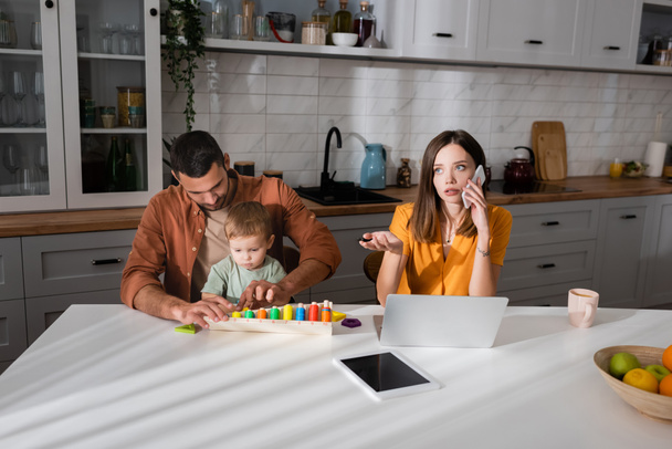Young freelancer talking on smartphone near gadgets and family playing game in kitchen  - 写真・画像