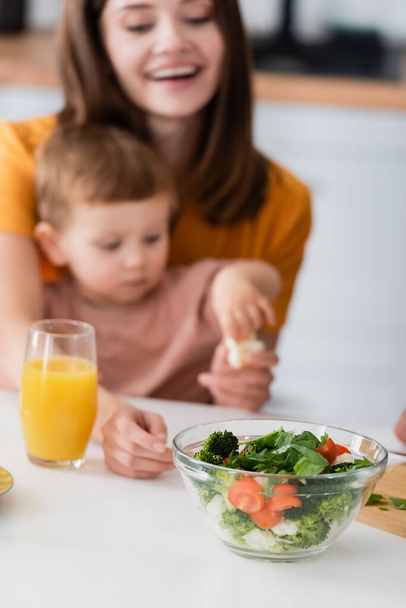 Fresh salad near orange juice and blurred mother and child at home  - Photo, Image