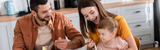 Young woman feeding son with pasta near husband at home, banner  - 写真・画像
