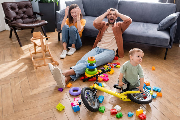 Kid standing near bike, building blocks and parents in living room  - Photo, image