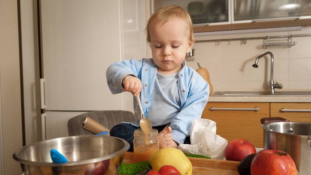 Pikkupoika istuu keittiön pöydällä ja syö hedelmäsosetta lusikalla. Käsitys terveellisestä ravitsemuksesta, ruokavaliosta ja lasten ruoasta - Valokuva, kuva