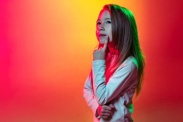 Half-length portrait of little thoughtful girl dreaming isolated on mangenta yellow studio background in neon light, filter. - Фото, зображення