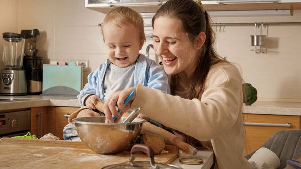 台所で生地や焼き菓子を作っている間、幸せな笑顔の母親は彼女の小さな赤ちゃんの息子と遊んで楽しんでいます。小さなシェフ、子供たち料理、健康的な栄養の概念. - 写真・画像