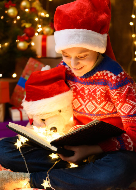 Child girl reading a book in new year or christmas decoration. She's wearing a red sweater and a Santa helper hat. Christmas tree decorated with toys. - Photo, image