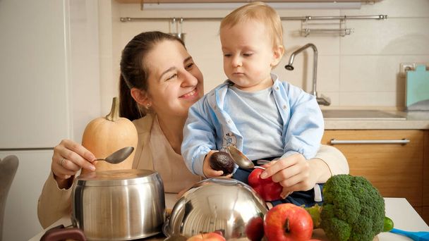 Bonne mère souriante avec bébé fils jouant avec des cuillères sur des casseroles dans la cuisine comme sur des tambours. Concept de petit chef, cuisine pour enfants, bon moment en famille ensemble. - Photo, image