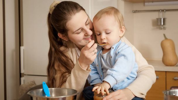 Smiling woman giving fruit puree to her baby son playing on kitchen. Concept of little chef, children cooking food, good family time together. - Zdjęcie, obraz