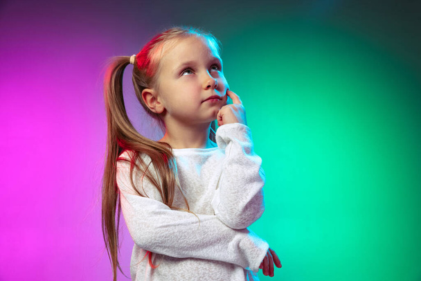 Half-length portrait of little thoughtful girl dreaming isolated on blue purple studio background in neon light, filter. - Photo, image