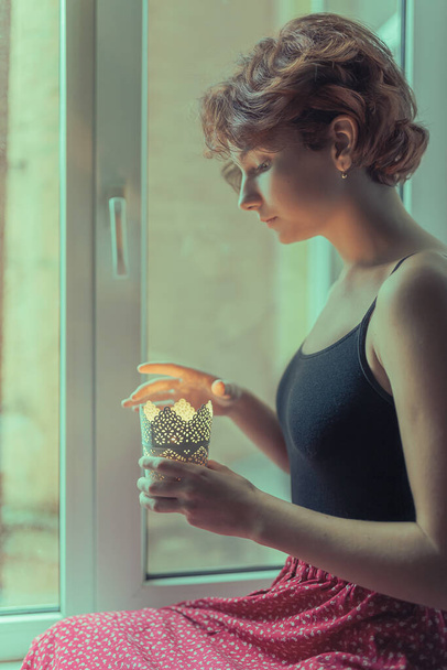 fiatal lány barna göndör haj karcsú alak ül az ablakpárkányon, és tartja égő gyertya a kezében. Magány, a találkozás és a várakozás reménye - Fotó, kép