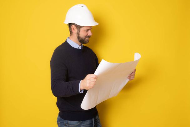 Jonge pensive architect man dragen aannemer helm holding project papier plan over geïsoleerde gele achtergrond - Foto, afbeelding