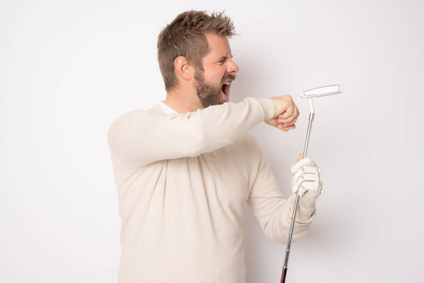 Νεαρός όμορφος άνδρας παίζει γκολφ με θυμωμένο πρόσωπο στέκεται απομονωμένος πάνω από λευκό φόντο. - Φωτογραφία, εικόνα