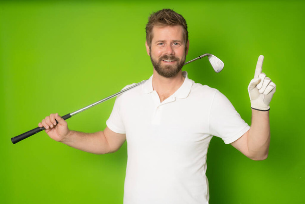 Guapo joven con camiseta blanca jugando al golf apuntando con el dedo hacia la cámara sobre un fondo verde aislado. - Foto, imagen