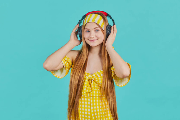 Studio portrait of Funny young blonde woman in yellow dress isolated on blue background. Girl Listen music with wireless headphones - Photo, Image