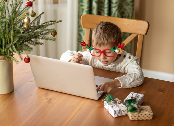  Pieni poika valkoisessa puserossa ja hauskoissa laseissa istuu kannettavan tietokoneen ääressä ja kirjoittaa kirjeen joulupukille. - Valokuva, kuva