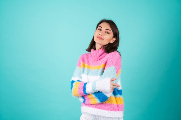 A young woman in a bright multicolored sweater on a blue background smiles cutely, looks at the camera, arms crossed on her chest, radiates calmness and confidence - Foto, afbeelding