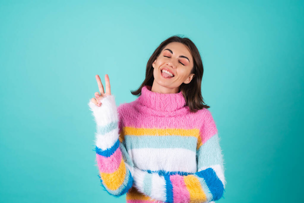 Fiatal nő egy fényes színes pulóver egy kék háttér aranyos mosolygós, játékos, bemutató nyelv és v gesztus - Fotó, kép