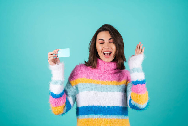 Young woman in a bright multicolored sweater on a blue background shows a credit card with a smile, excited, surprised - Photo, Image