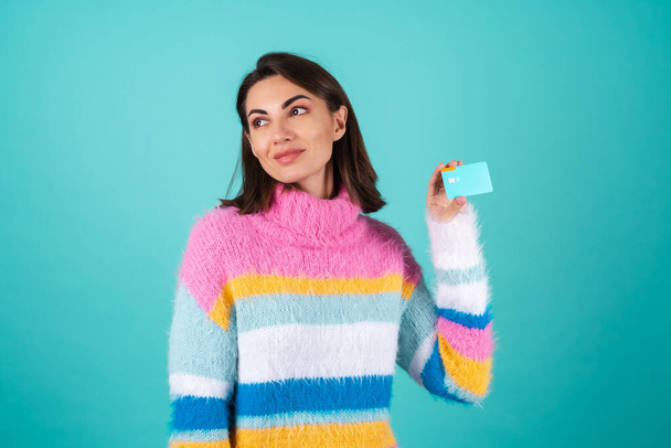 Young woman in a bright multicolored sweater on a blue background shows a credit card with a smile - Фото, зображення