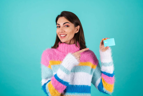 Jovem mulher em uma camisola multicolorida brilhante em um fundo azul mostra um cartão de crédito com um sorriso - Foto, Imagem