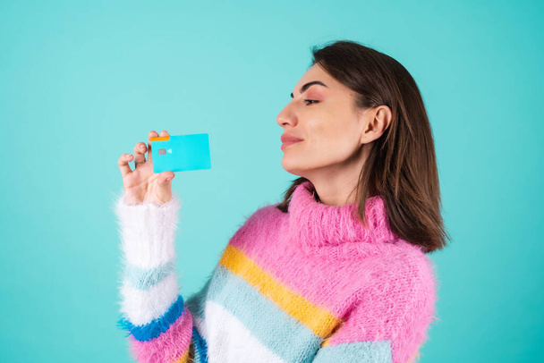 Uma jovem mulher em uma camisola multicolorida brilhante em um fundo azul mostra um cartão de crédito com um espaço vazio, sorrisos alegremente, bela maquiagem - Foto, Imagem
