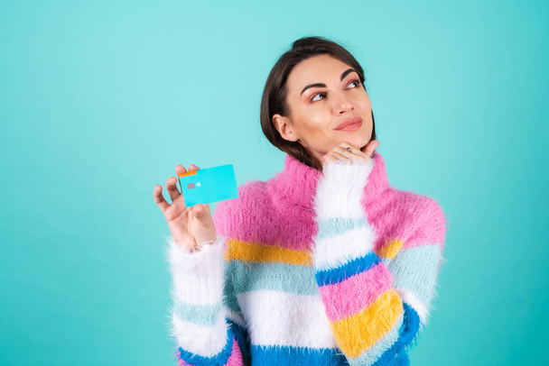 Una mujer joven en un suéter multicolor brillante sobre un fondo azul muestra una tarjeta de crédito, mira pensativamente, soñadoramente a un lado, con una sonrisa en su cara - Foto, Imagen