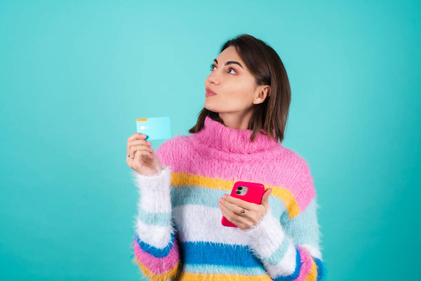 Mujer joven en un suéter multicolor brillante sobre un fondo azul sostiene una tarjeta de crédito, paga por las compras en línea en un teléfono móvil, mira pensativamente a un lado - Foto, imagen