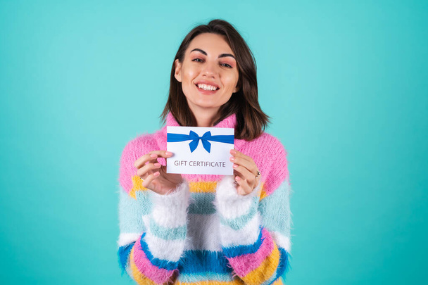 Young woman in a bright multicolored sweater on a blue background holding a gift certificate, cute smiling - Photo, Image