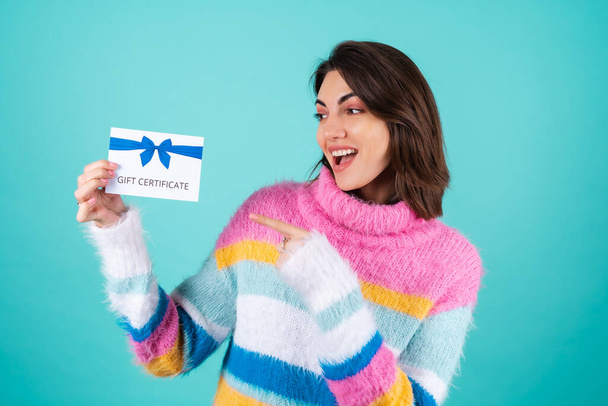 Giovane donna in un luminoso maglione multicolore su sfondo blu in possesso di un buono regalo, sorridente carino, che punta con un dito a un voucher - Foto, immagini