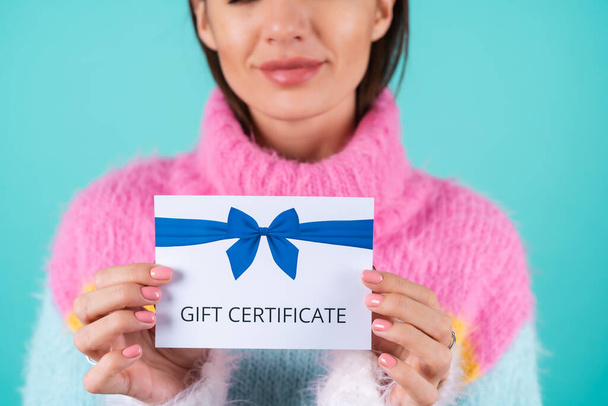 Young woman in a bright multicolored sweater on a blue background holds a gift certificate, smiles enthusiastically, beautiful makeup, nude plump lips - Photo, Image