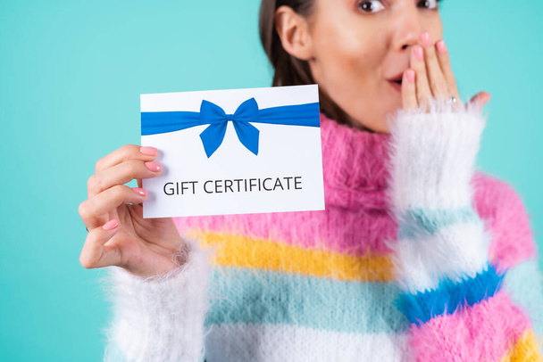 Mujer joven en un suéter multicolor brillante sobre un fondo azul tiene un certificado de regalo, sonríe con entusiasmo, hermoso maquillaje, labios rellenos desnudos - Foto, imagen