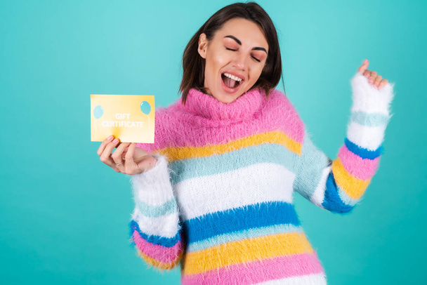 Young woman in a bright multicolored sweater on a blue background holds a gift certificate, enthusiastically jumping with happiness - Photo, Image