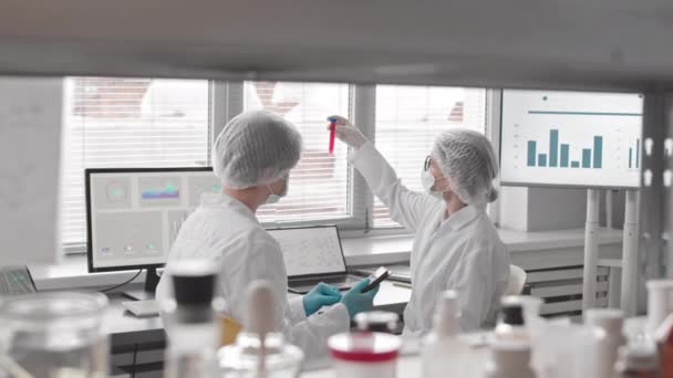 Середина двох кавказьких жінок-вчених, одягнених у маску обличчя, рукавички, медичні сукні і капелюхи сидять за столом у лабораторії вдень, вивчаючи пробірку, розмовляючи і використовуючи планшетний комп'ютер. - Кадри, відео