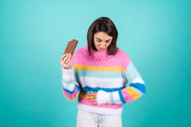 Young woman in a bright multicolored sweater on a blue background suffering from indigestion and abdominal pain, holding a bar of milk chocolate - Photo, Image