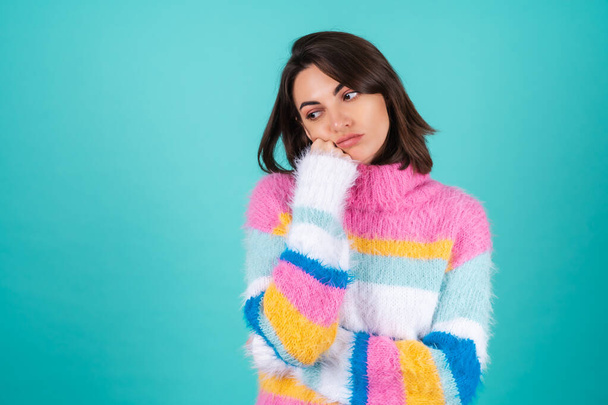 A young woman in a bright multicolored sweater on a blue background stands with a bored indifferent look, a fist on her chin - Photo, Image