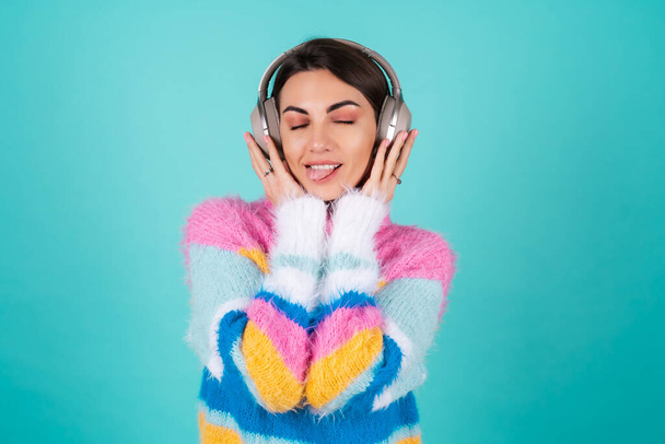 Young woman in a bright multicolored sweater on a blue background in large noise canceling headphones, enjoying her favorite music, music lover - 写真・画像